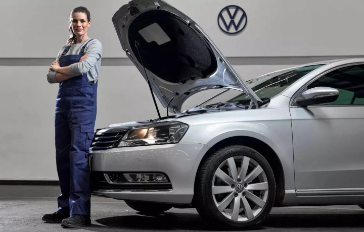 Un meccanico in tuta blu sta in piedi con sicurezza, con le braccia incrociate, accanto a una Volkswagen argentata, con il cofano aperto, a simboleggiare la fiducia e la competenza che spingono i clienti a scegliere Caltabiano auto.